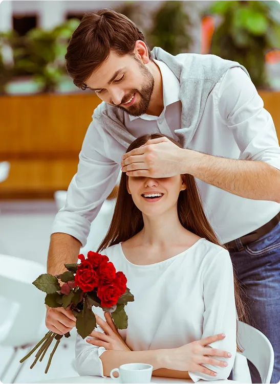 A smiling man surprises a woman by covering her eyes as she holds a bouquet of red roses